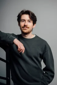 Casual portrait of a man with brown hair and light beard, wearing a dark green sweatshirt, resting his forearm on a railing against a gray backdrop.