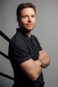 Portrait of a man with short brown hair and stubble, wearing a black polo, arms crossed, against a gray studio backdrop.
