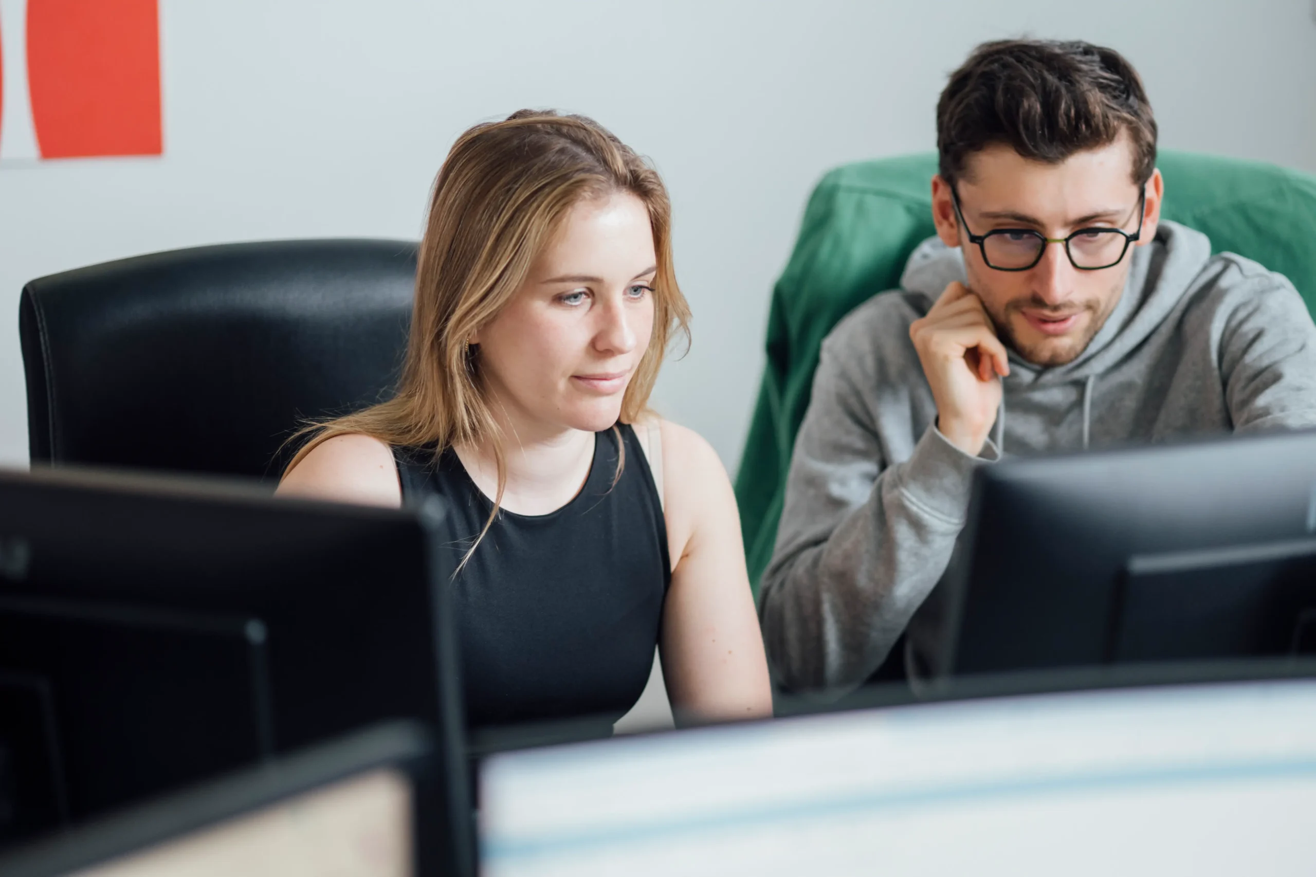 Two coworkers sit side by side at computer monitors, focused on their screens in a bright office.