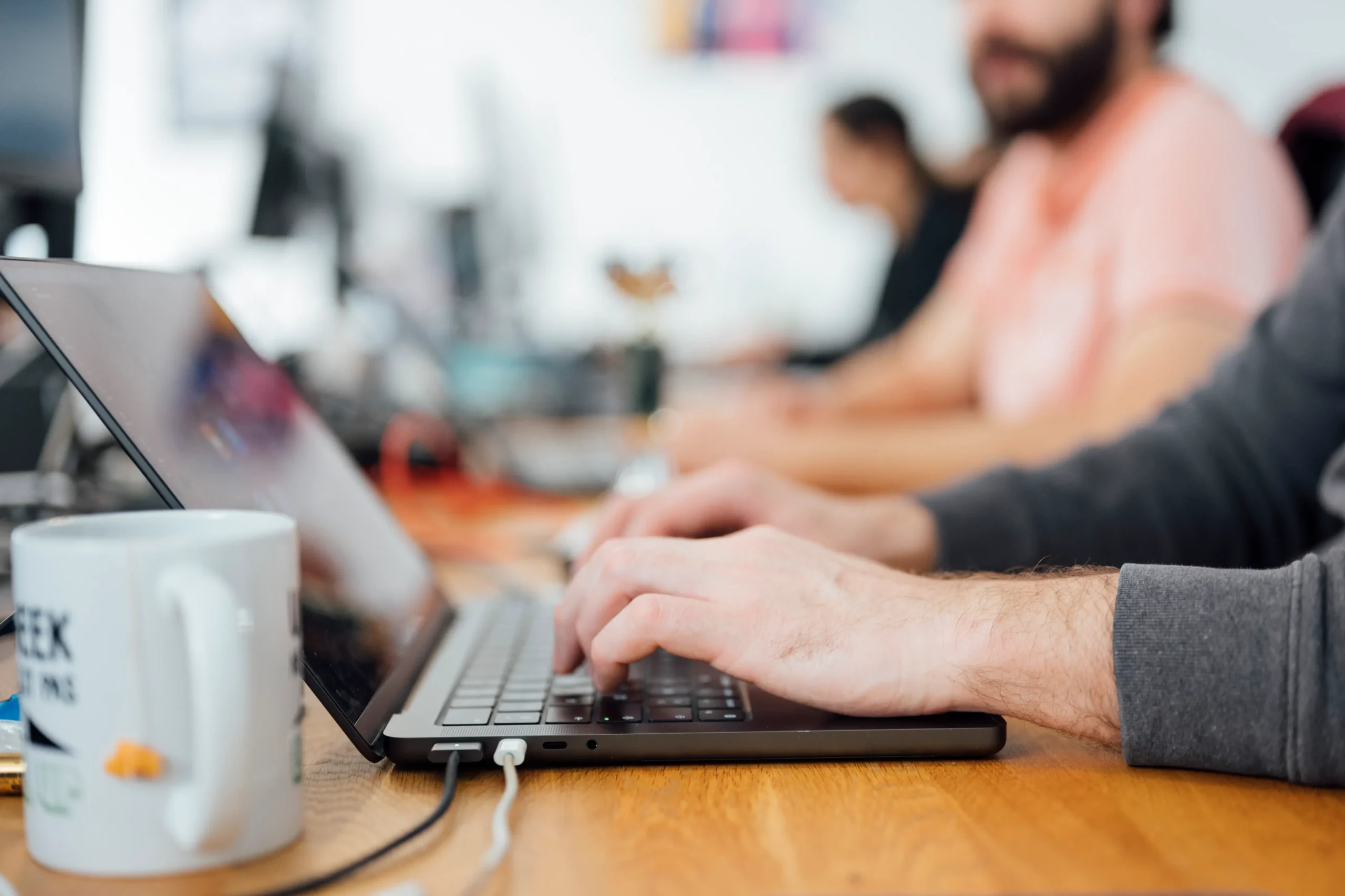People typing on laptops at a wooden table in a bright office, foreground mug visible.