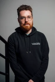 Portrait of a man with curly brown hair and a red beard wearing round glasses and a black hoodie against a gray backdrop