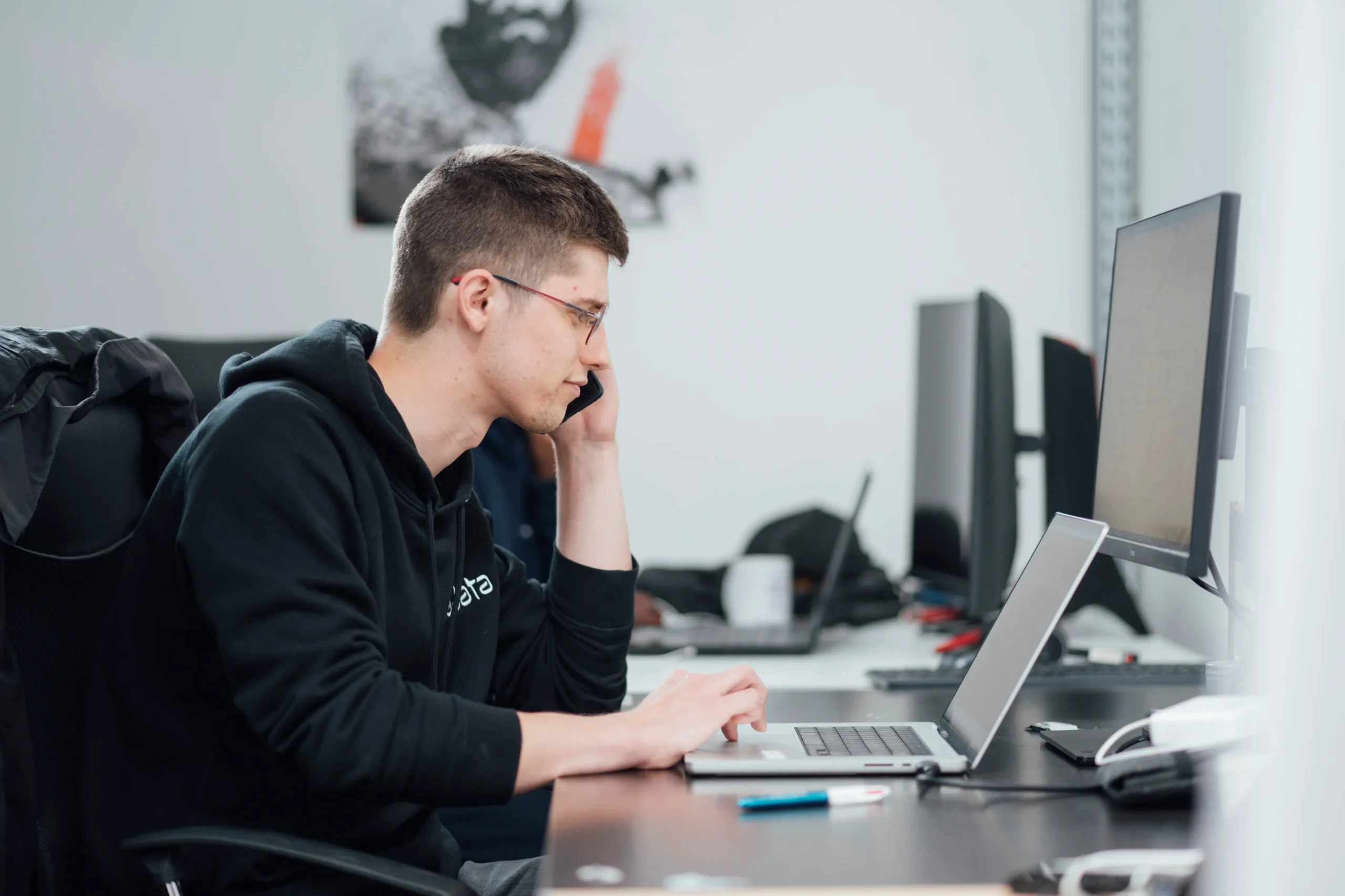 Man in a black hoodie sits at a desk, talking on a phone while typing on a laptop in a bright office workspace.